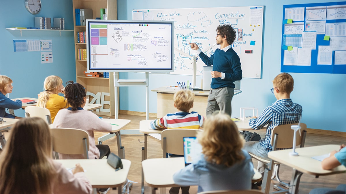 salle de classe dans une école, il y a des jeunes élèves face à un tableau connecté montré par l'enseignant.
